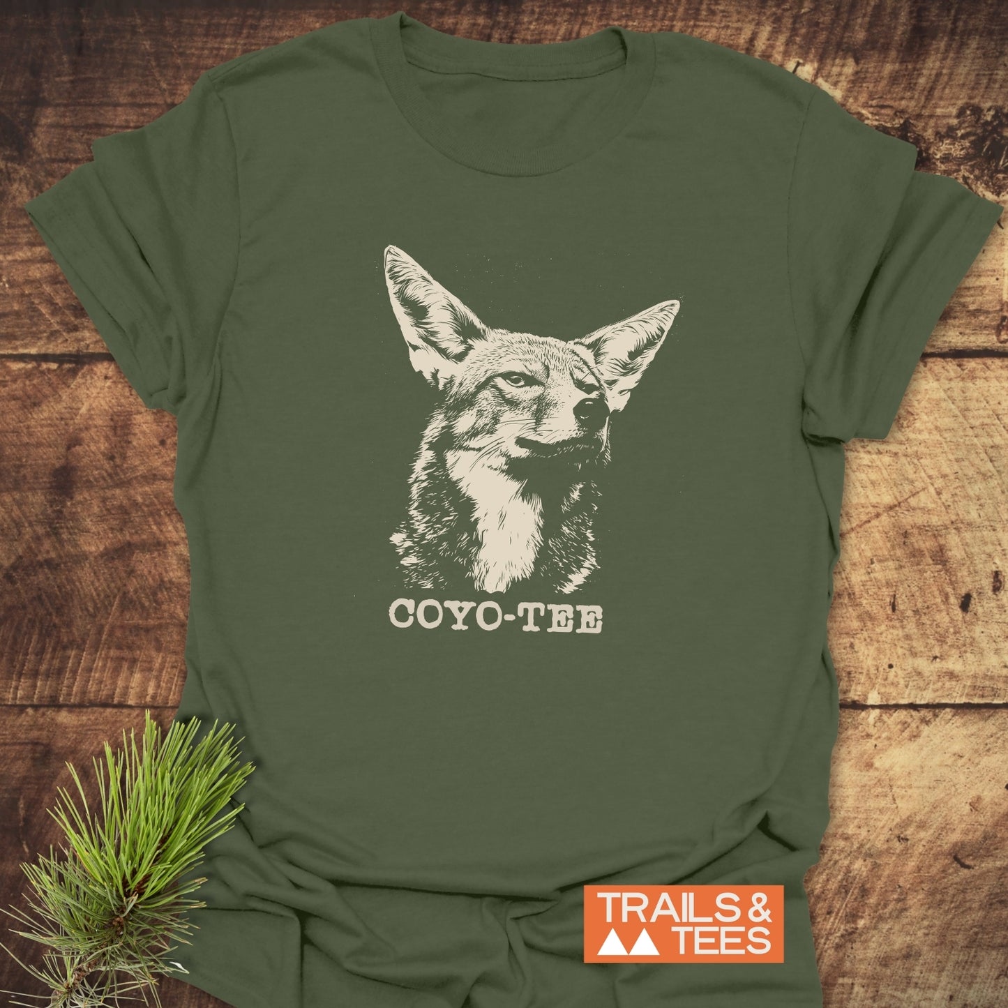 Forest-green Coyo-Tee T-Shirt with a white coyote illustration and pun caption, laid on wood. Includes TRAILS & TEES label and pine sprig accent.