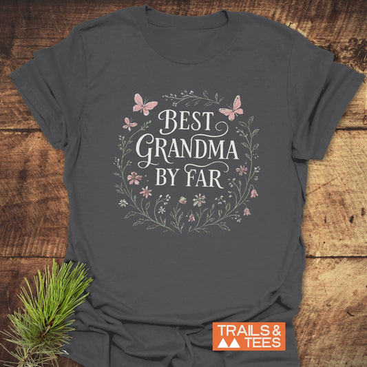 Charcoal-gray Best Grandma Flower T-Shirt with a floral wreath and butterfly design, highlighting comfort and durability, laid on a wooden surface with TRAILS & TEES logo.