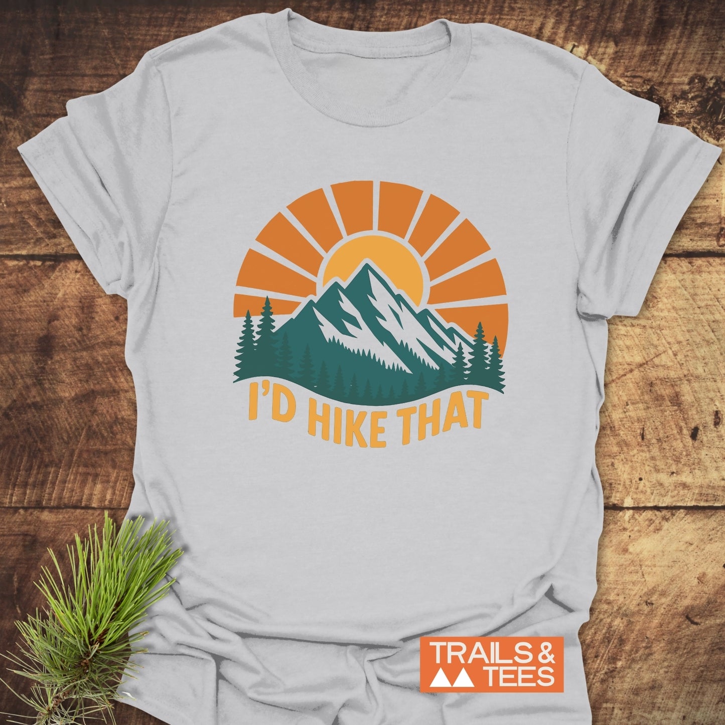 I'd Hike That T-Shirt with mountain graphic, pine trees, and slogan. Soft ring-spun cotton, durable print. Trails & Tees logo visible, pine needles accentuate theme.