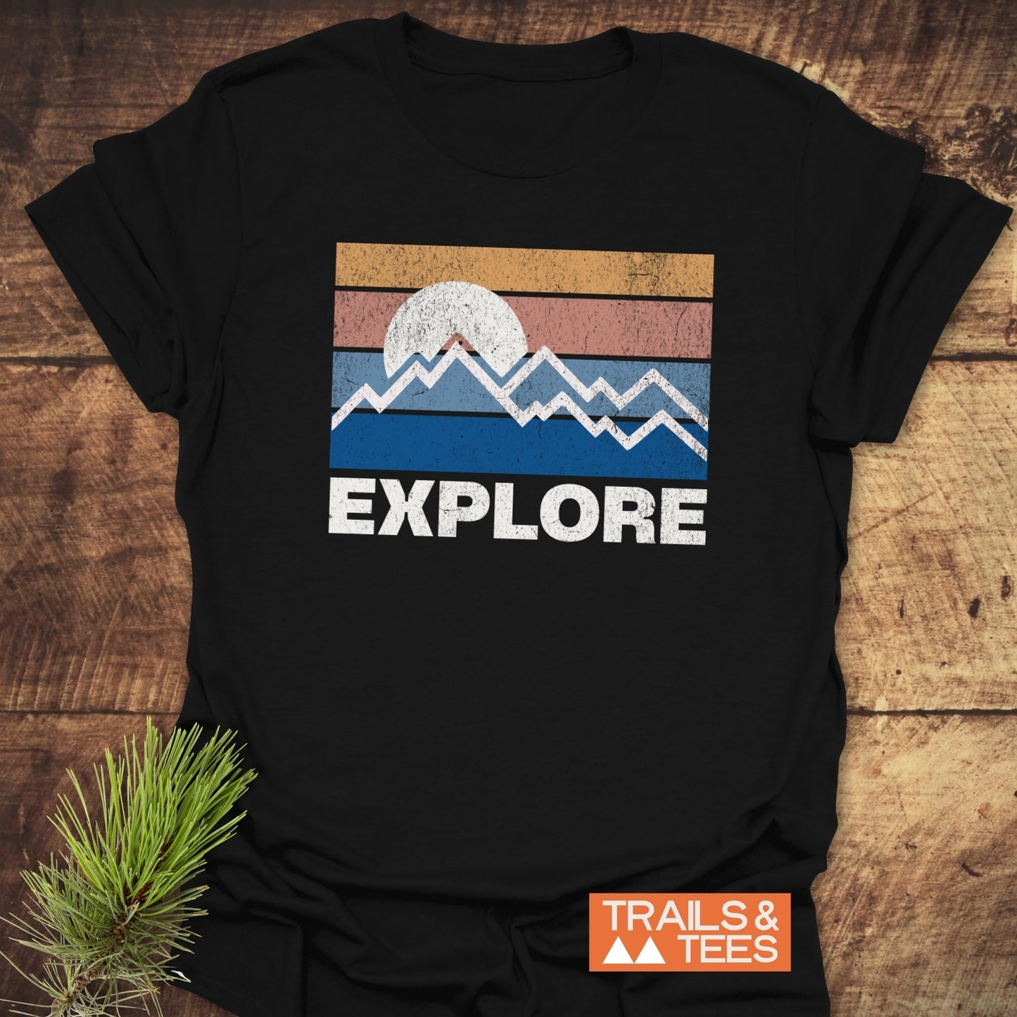 Explore Retro Sunset T-Shirt with a mountain graphic and EXPLORE text, laid flat with a pine sprig, showcasing outdoor-inspired design and comfort.