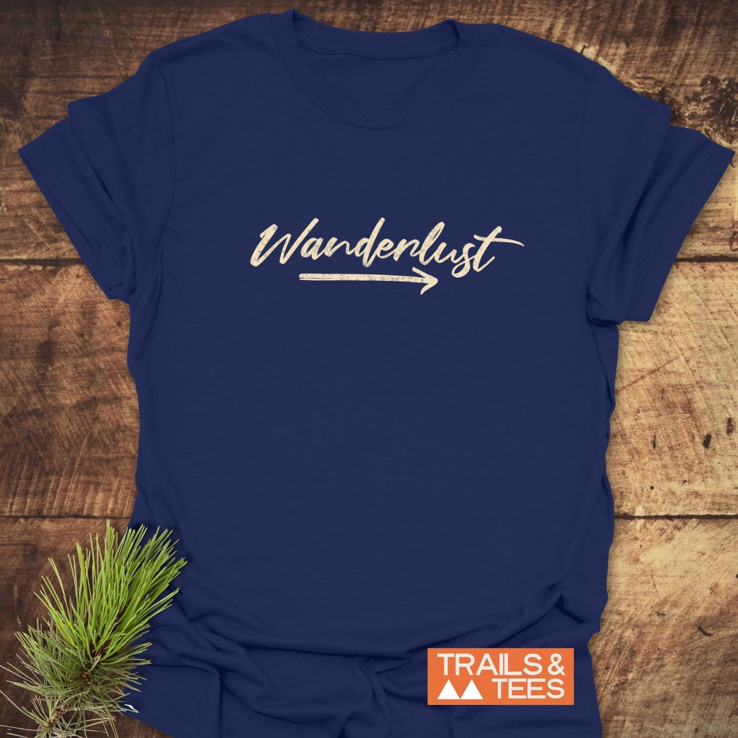 Wanderlust T-Shirt in navy with Wanderlust script and arrow design, featuring soft ring-spun cotton for comfort, displayed with pine sprig and Trails & Tees logo.