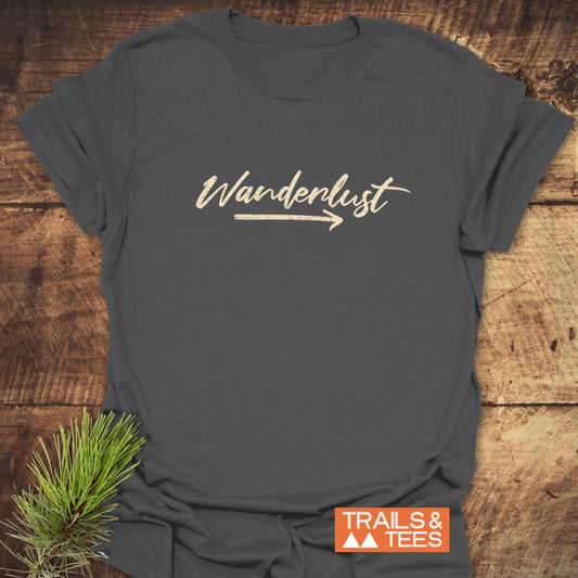 Wanderlust T-Shirt: Charcoal-gray crew-neck with Wanderlust text and arrow, showcasing comfort and durability. Features Trails & Tees logo, pine sprig accent.