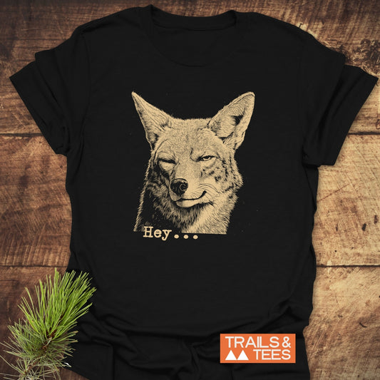 Hey... Coyote Funny T-Shirt featuring a sketched winking coyote and “Hey…” text, styled on wood with a pine sprig, Trails & Tees logo visible.