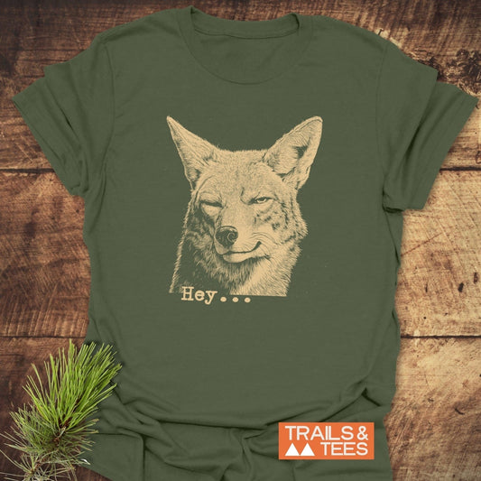 Hey... Coyote Funny T-Shirt featuring a winking coyote illustration and Hey... text, styled with a pine sprig, showcases outdoor-inspired charm on a soft, pre-shrunk, ring-spun cotton fabric.