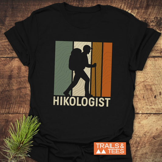 Hikologist T-Shirt featuring a hiker silhouette over colored blocks, with HIKOLOGIST text; crafted from soft cotton for outdoor enthusiasts, includes TRAILS & TEES logo.