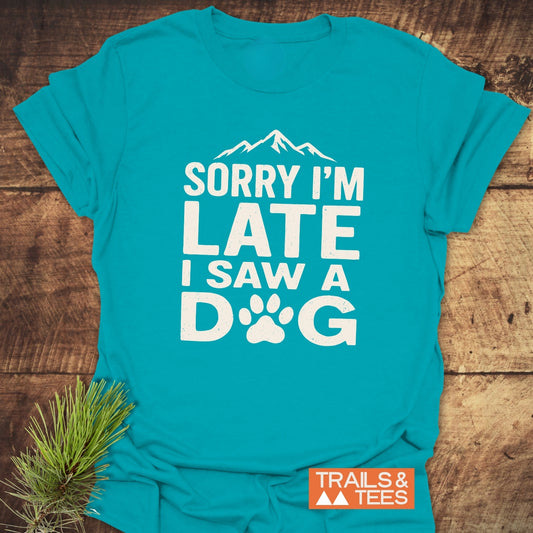 I Saw A Dog T-Shirt with mountain graphic, paw-print, and SORRY I'M LATE slogan, laid flat on wood, Trails & Tees logo visible.