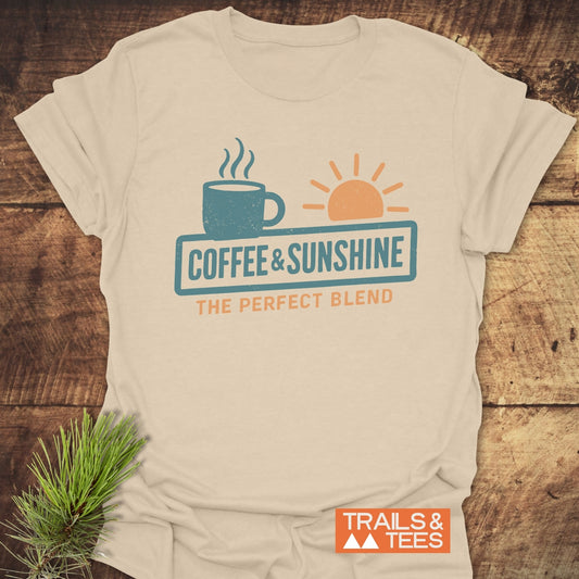 Coffee & Sunshine T-Shirt with teal and orange mug and sun graphic, laid on wood. Soft, lightweight cotton, pre-shrunk, durable print, designed for outdoor enthusiasts.
