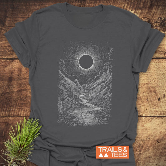 Eclipse Canyon T-Shirt featuring a detailed mountain valley and celestial illustration on dark-gray cotton, laid flat with a pine sprig accent.