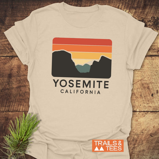 Retro Yosemite T-Shirt featuring a mountain valley silhouette on a sunset design, laid on wood with pine sprig, crafted for comfort and adventure.
