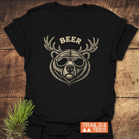Beer Funny T-Shirt featuring a bear with sunglasses and antlers graphic. Laid on wood with pine sprigs, highlighting its outdoor, rustic theme.