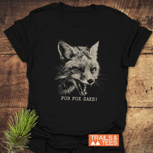 For Fox Sake! Funny T-Shirt featuring a detailed fox illustration and slogan, laid on wood; Trails & Tees logo and pine sprig for styling.
