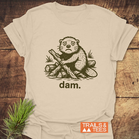 Beaver - Dam Funny T-Shirt featuring a beaver illustration stacking logs with dam text, showcasing comfort and durability. Laid on wood with pine sprig.