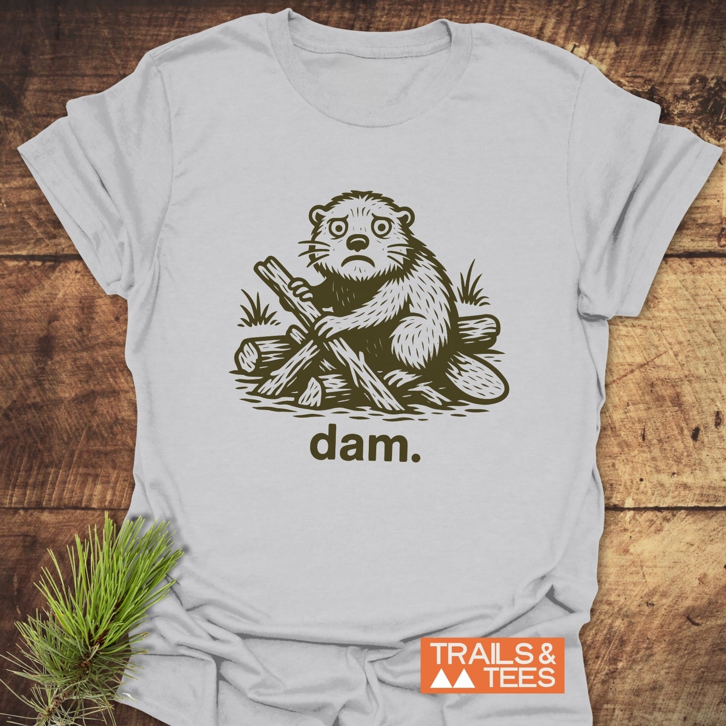 Light-gray T-shirt featuring a forlorn cartoon beaver with logs, “dam.” text, and “TRAILS & TEES” logo, highlighting comfort and outdoor adventure appeal.