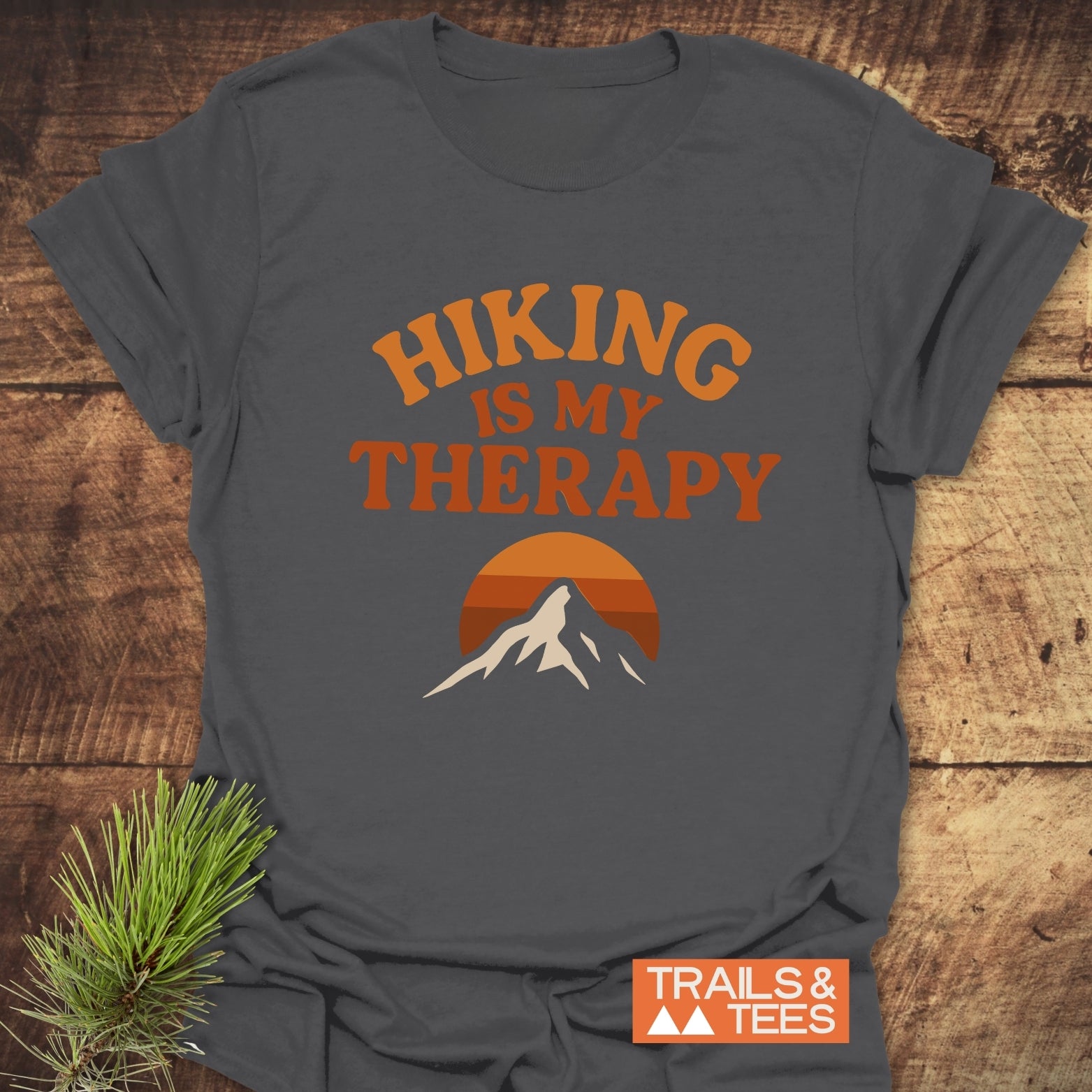 Hiking Therapy T-Shirt with HIKING IS MY THERAPY and mountain graphic, made of soft ring-spun cotton, laid flat on wood surface with pine sprig accent.
