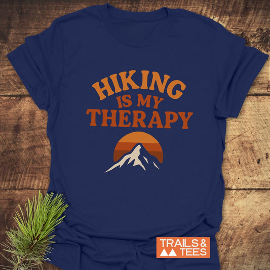 Navy-blue Hiking Therapy T-Shirt with a mountain sunset graphic and slogan, laid on wood. Crafted from soft cotton, it offers comfort and durability.
