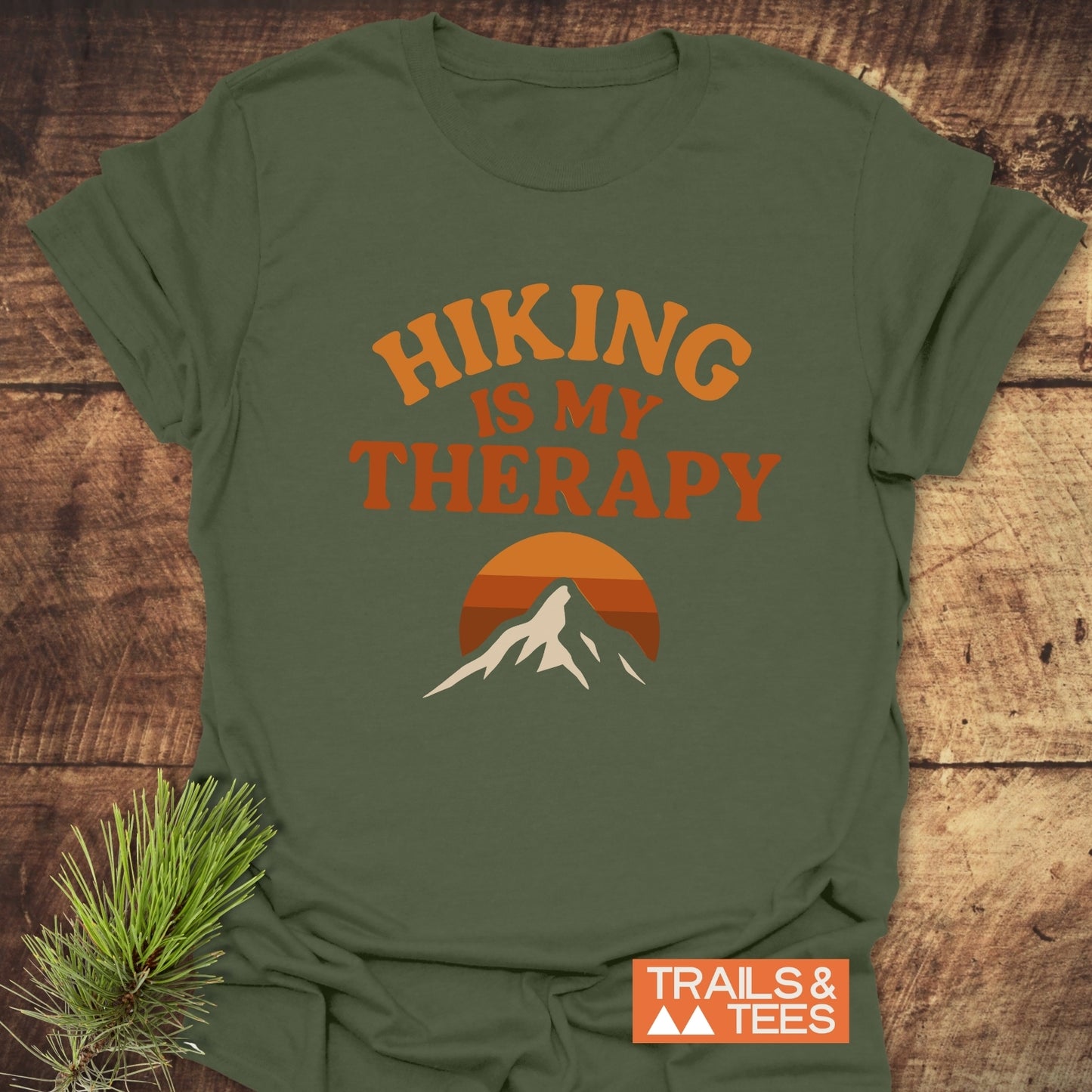 Hiking Therapy T-Shirt with HIKING IS MY THERAPY slogan, mountain silhouette, printed on soft cotton. Laid flat with pine sprigs for an outdoorsy vibe.