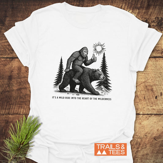 Wild Ride Bigfoot T-Shirt features a sasquatch on a bear, thumbs-up, with pine trees and sun, slogan beneath, showcasing outdoor adventure spirit.