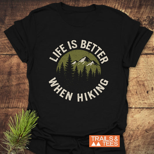 Life Is Better When Hiking T-Shirt featuring a circular evergreen and mountain graphic, laid on wood with a pine branch, showcasing outdoor adventure spirit.