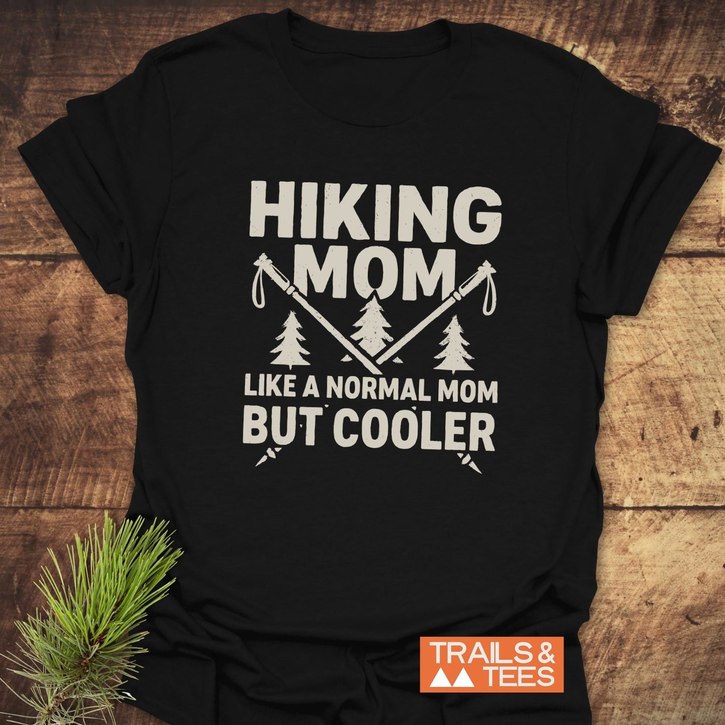 Hiking Mom T-Shirt features crossed hiking poles and pine trees, with text highlighting a cool mom theme. Crafted from lightweight cotton with durable prints.