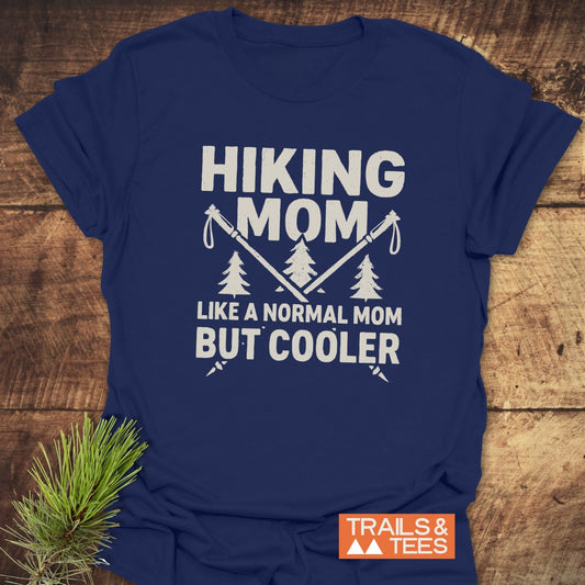 Navy-blue Hiking Mom T-shirt with slogan, evergreen trees, and hiking poles. Made from soft cotton, pre-shrunk, durable prints. Laid on wooden surface.
