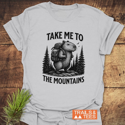Capybara Mountains T-Shirt featuring a hiking capybara illustration with TAKE ME TO THE MOUNTAINS, crafted for comfort with durable print and pre-shrunk fit.