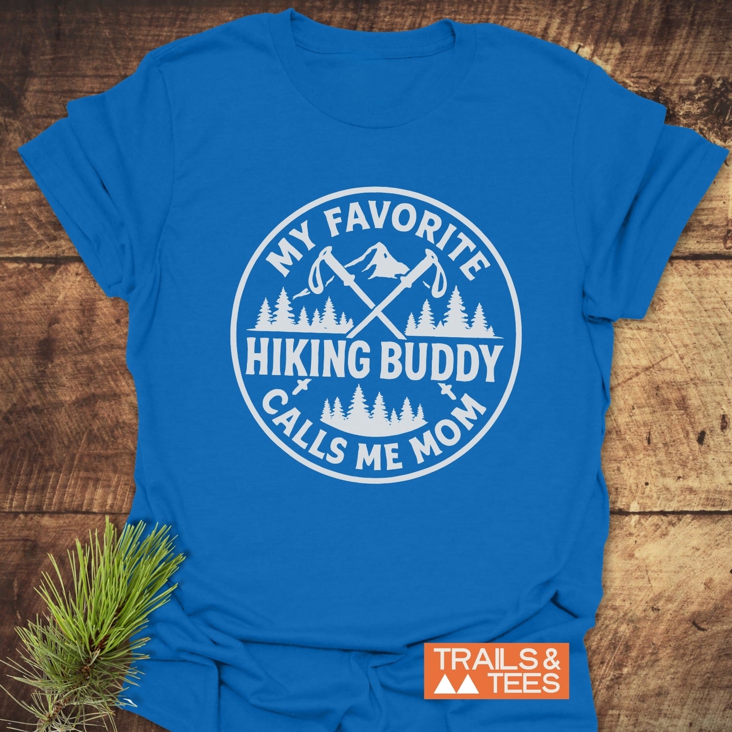 Hiking Buddy Mom T-Shirt featuring a white graphic of hiking poles, pine trees, and a mountain. Laid flat with a pine sprig, showcasing outdoor-inspired design.