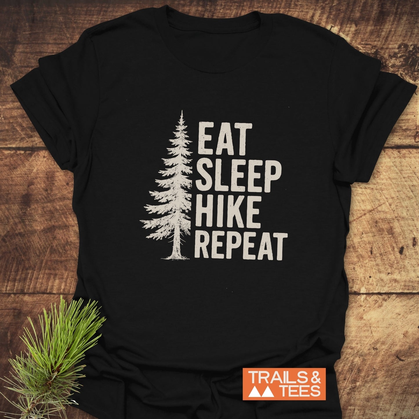 Hike, Repeat T-Shirt with white pine tree graphic and text EAT SLEEP HIKE REPEAT on black cotton. Features small TRAILS & TEES. logo.