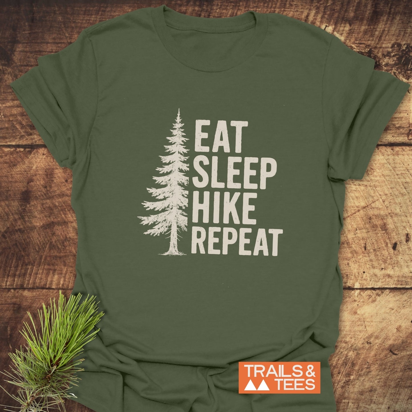 Hike, Repeat T-Shirt featuring a white pine tree silhouette and text on olive-green cotton, styled with a pine sprig. Durable, pre-shrunk, and sustainably printed.