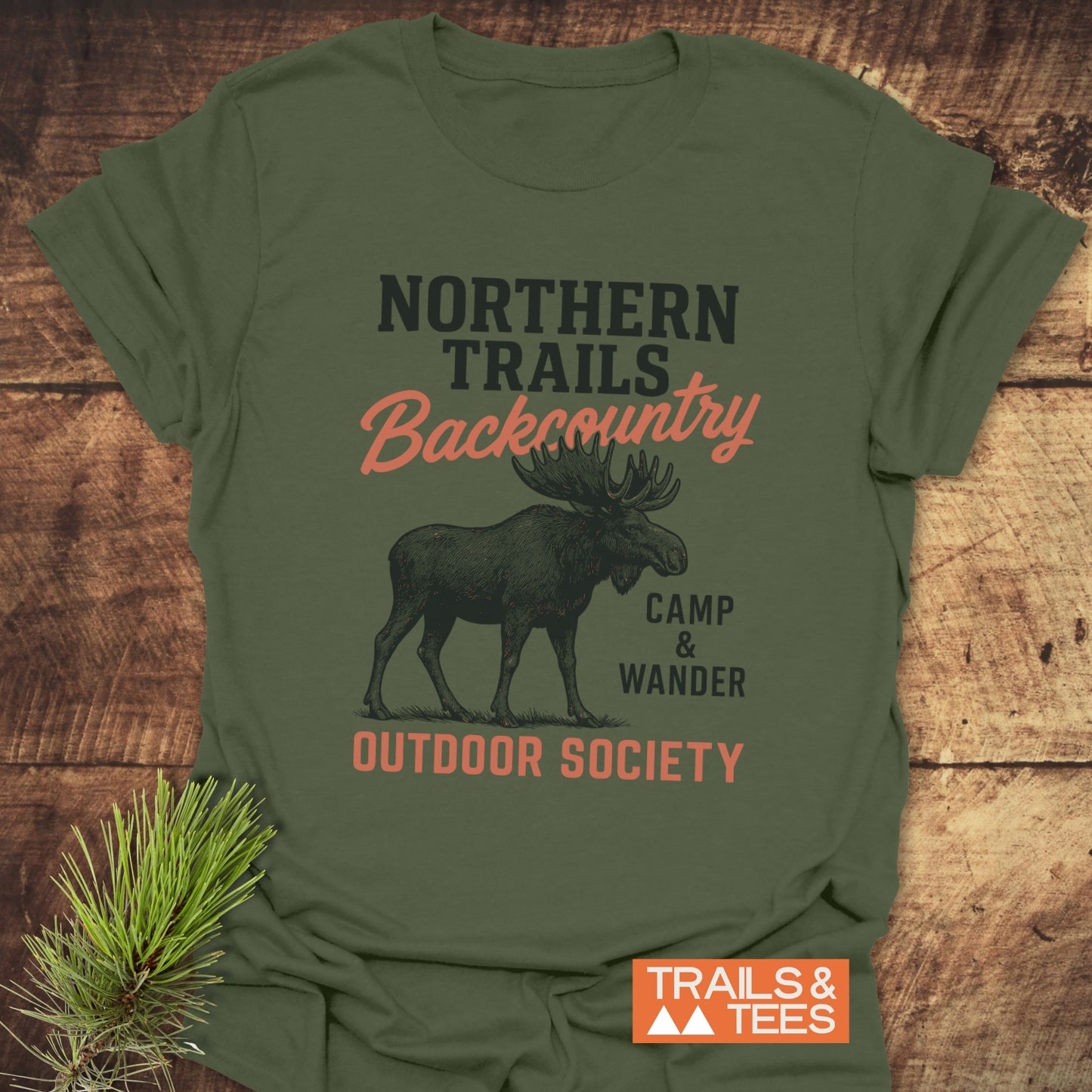 Northern Trails Moose T-Shirt featuring a vintage moose graphic on olive-green fabric, emphasizing outdoor adventure. Laid on rustic wood with pine sprig and Trails & Tees label.