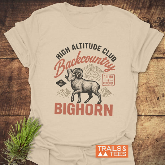 High Altitude Club T-Shirt featuring a ram graphic, mountain outlines, and text details, laid on wood. Lightweight, durable, and pre-shrunk for comfort.