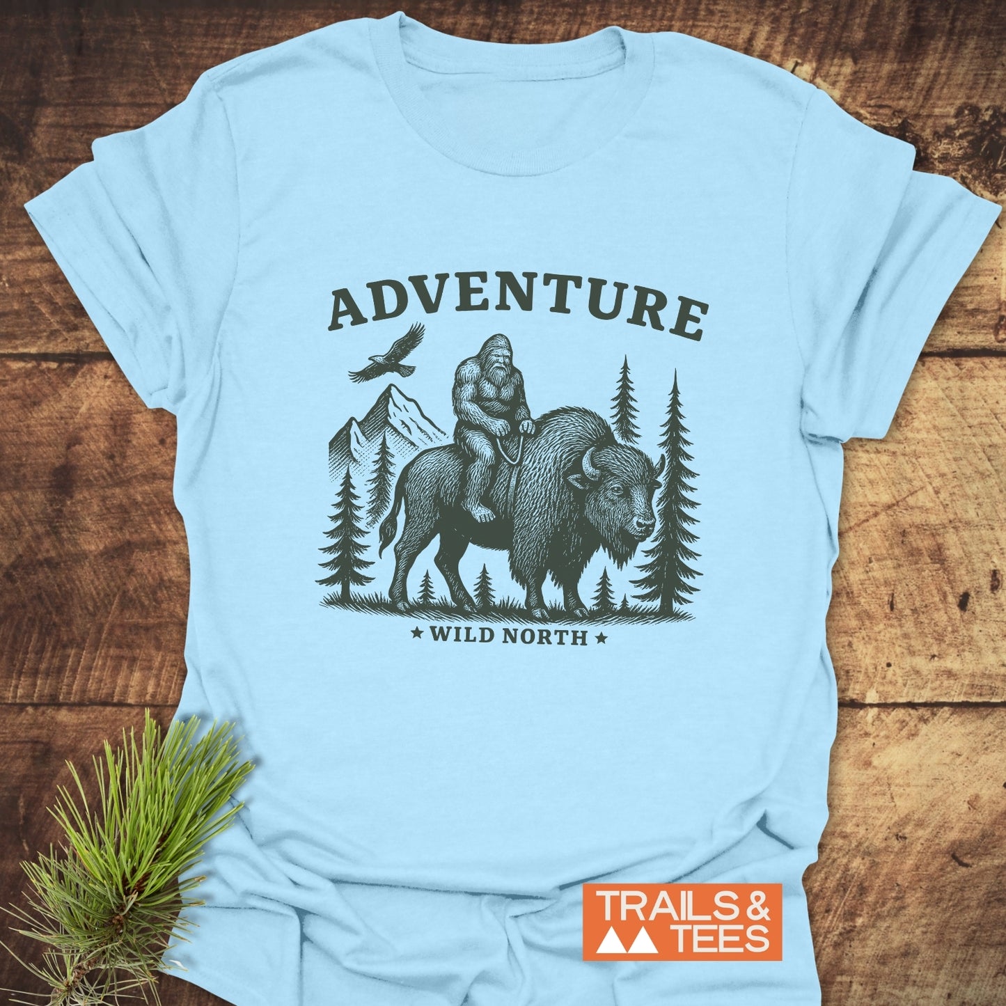 Bigfoot & Bison T-Shirt featuring Sasquatch riding a bison in a wilderness scene, with ADVENTURE and WILD NORTH text, ideal for outdoor enthusiasts.