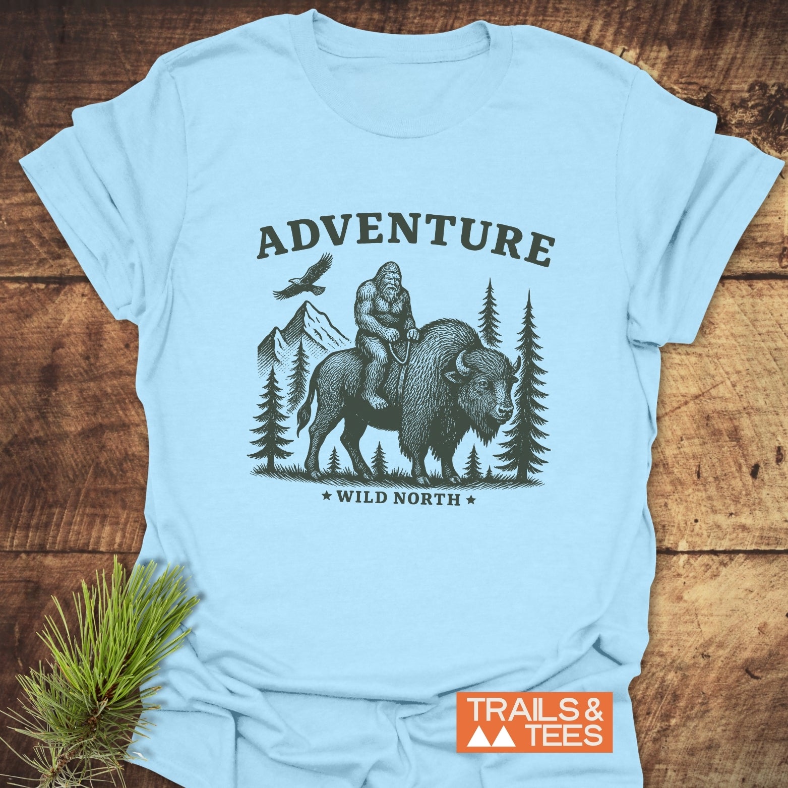 Bigfoot & Bison T-Shirt featuring Sasquatch riding a bison in a wilderness scene, with ADVENTURE and WILD NORTH text, ideal for outdoor enthusiasts.