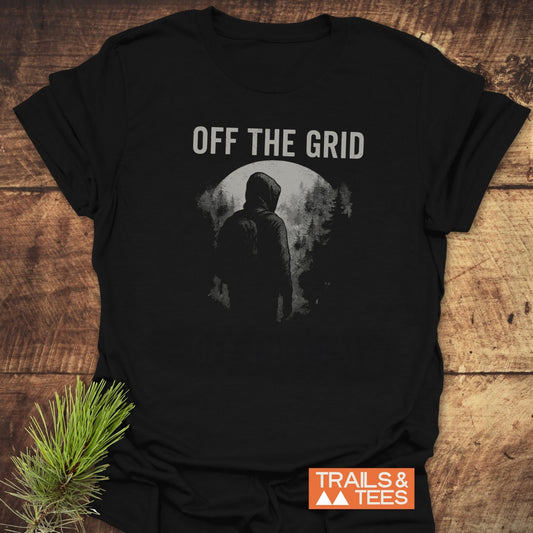 Off The Grid T-Shirt featuring a hooded figure graphic under OFF THE GRID text, laid flat on wood with Trails & Tees logo visible.