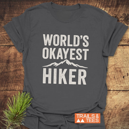 Dark gray World's Okayest Hiker T-shirt with white print and mountain graphic, laid flat. Features soft, lightweight cotton, durable print, and eco-friendly production.