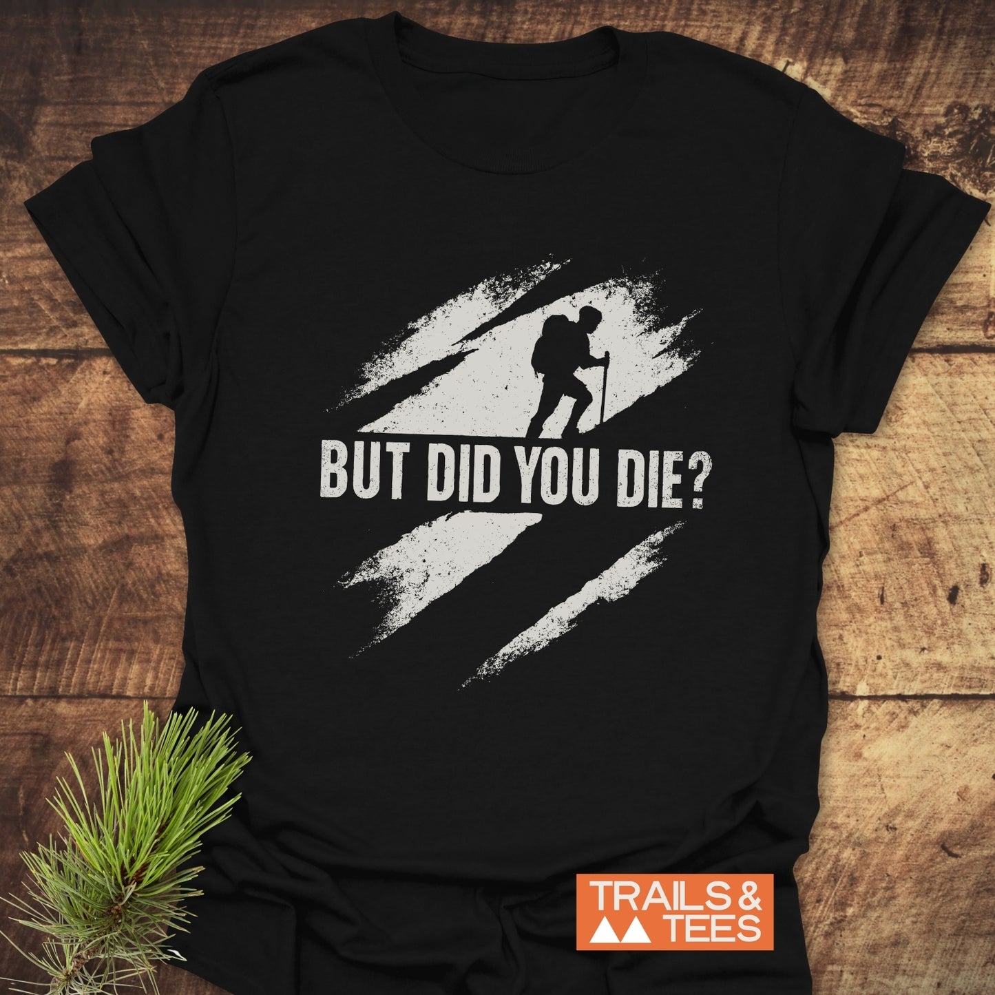 But Did You Die? Hiking T-Shirt with a claw-mark graphic and TRAILS & TEES logo, showcasing a backpacker silhouette, laid on a wooden surface.
