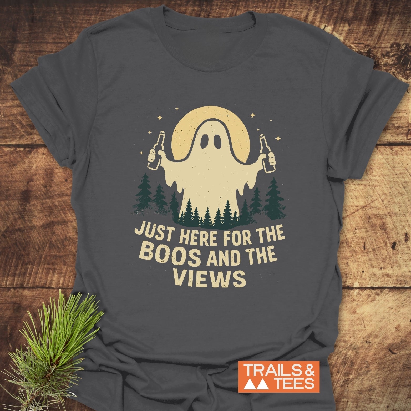 Boos And Views Halloween T-Shirt featuring a ghost with beers, moon, and trees. Laid on wood, showcasing Trails & Tees logo.