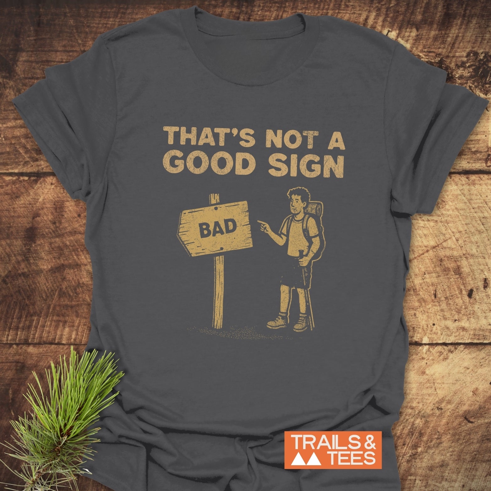 Bad Sign Funny T-Shirt: Charcoal-gray tee with a distressed yellow-tan print showing a hiker pointing at a crooked sign. Soft, lightweight, ring-spun cotton.