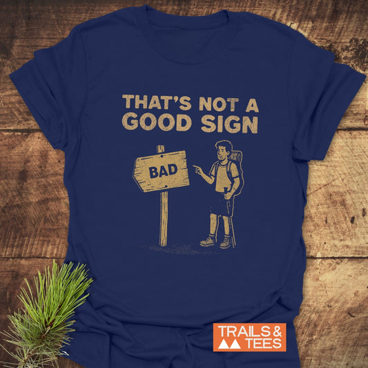 Bad Sign Funny T-Shirt with cartoon hiker and THAT’S NOT A GOOD SIGN distressed tan print, ideal for outdoor enthusiasts, laid flat on wooden surface.