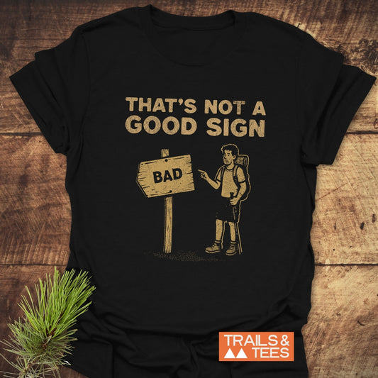 Bad Sign Funny T-Shirt featuring a cartoon hiker with a trail sign, made of soft cotton for outdoor enthusiasts. Includes a small TRAILS & TEES logo.