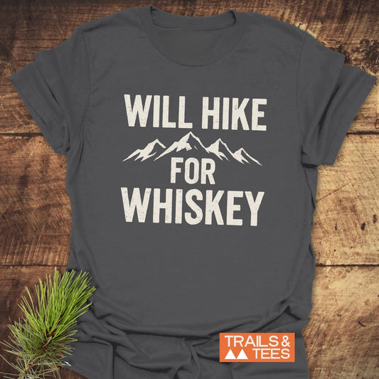 Will Hike For Whiskey T-Shirt folded on wood, featuring mountain-range graphic and slogan. Includes “TRAILS & TEES” label, embodying adventure-ready, soft cotton comfort.