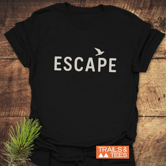 Escape Minimal T-Shirt featuring ESCAPE and bird design on black fabric; laid on wood with pine sprig, showcasing comfort and outdoor inspiration.