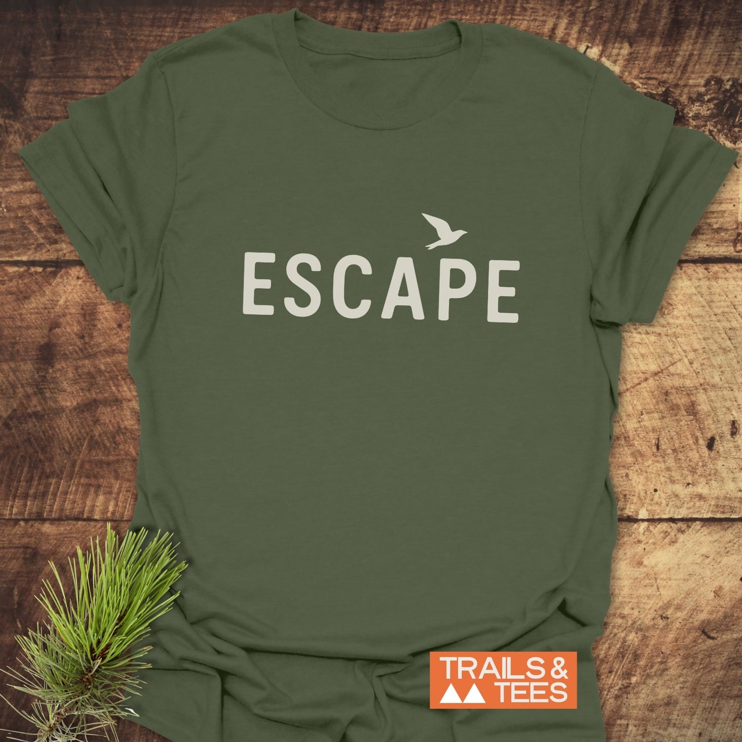 Escape Minimal T-Shirt in olive-green, featuring ESCAPE text with a bird silhouette. Laid on wood with pine needles and TRAILS & TEES logo visible.