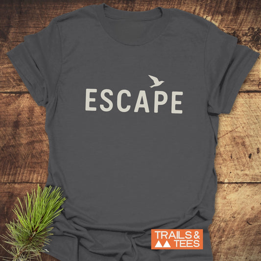 Escape Minimal T-Shirt featuring ESCAPE with a bird silhouette on dark-gray fabric, laid on rustic wood, styled with pine, and Trails & Tees logo.