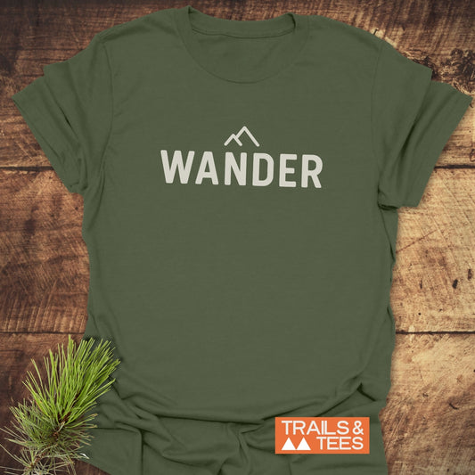 Olive-green 547 Wander Minimalist T-Shirt with mountain graphic and WANDER text, displayed on wood surface, highlighting its outdoorsy theme and Trails & Tees branding.