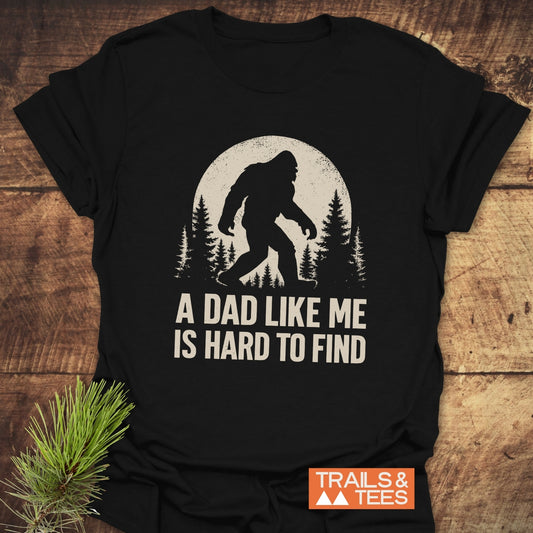 Vintage Bigfoot Dad T-Shirt with cream Bigfoot graphic and caption, A DAD LIKE ME IS HARD TO FIND, on black, laid flat, featuring Trails & Tees logo.