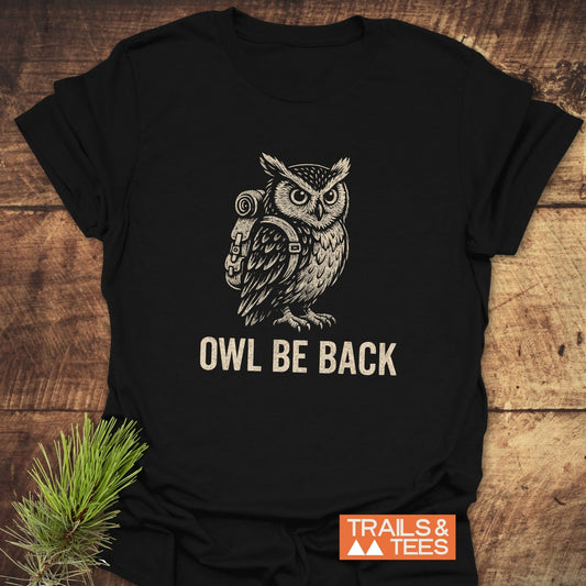 Owl Be Back Funny T-Shirt with an illustrated owl in a backpack and slogan, laid on wood. Durable, soft cotton for outdoor lovers, featuring Trails & Tees logo.