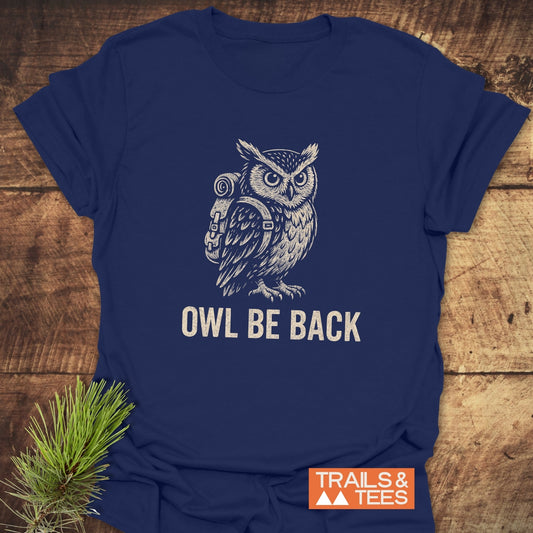 Navy-blue Owl Be Back Funny T-Shirt featuring a white owl with a hiking pack, “TRAILS & TEES” logo, pine bough accent, on wooden surface.