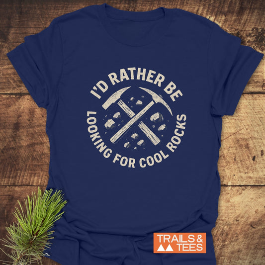 I'd Rather... Rocklover T-Shirt displaying crossed rock-hammers graphic, slogan, and 'TRAILS & TEES' logo, set on a wooden surface with pine sprig.