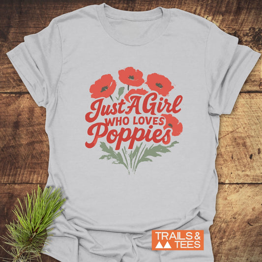 Girl Poppies T-Shirt featuring red poppy bouquet and text on light fabric, showcasing durable print and comfort, laid on rustic wood with pine sprig beside.
