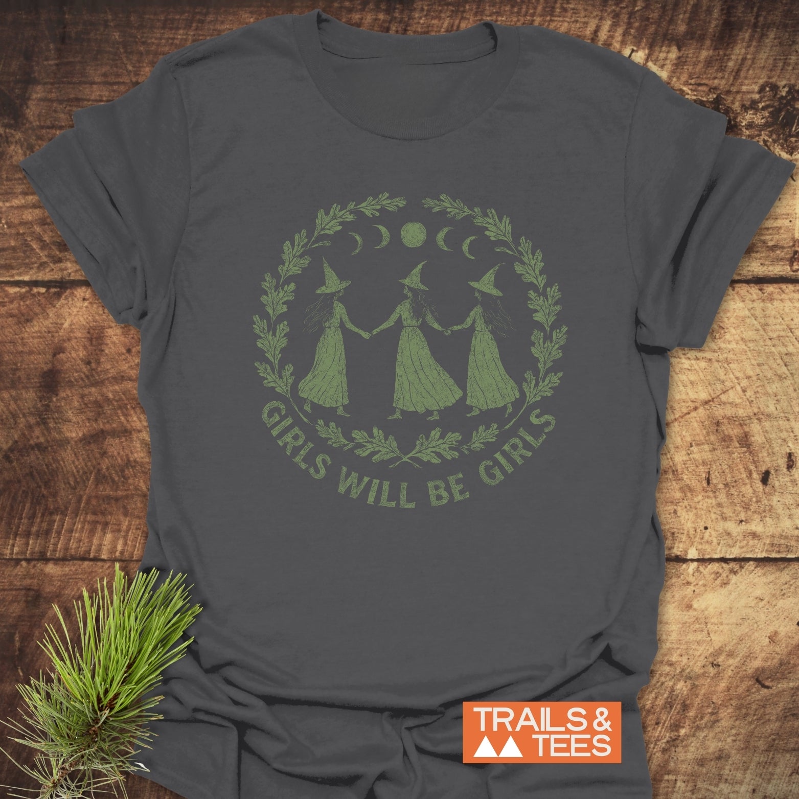 Witches Halloween T-Shirt featuring a graphic of witches with moon phases and slogan, on soft, durable cotton, styled with a pine sprig, by Trails & Tees.